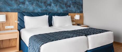 Un lit bien fait, avec du linge de lit blanc et une couvre-lit à motifs bleus et blancs, une table de chevet en bois avec une lampe, et un mur recouvert d’un papier peint à motifs de feuilles bleues.