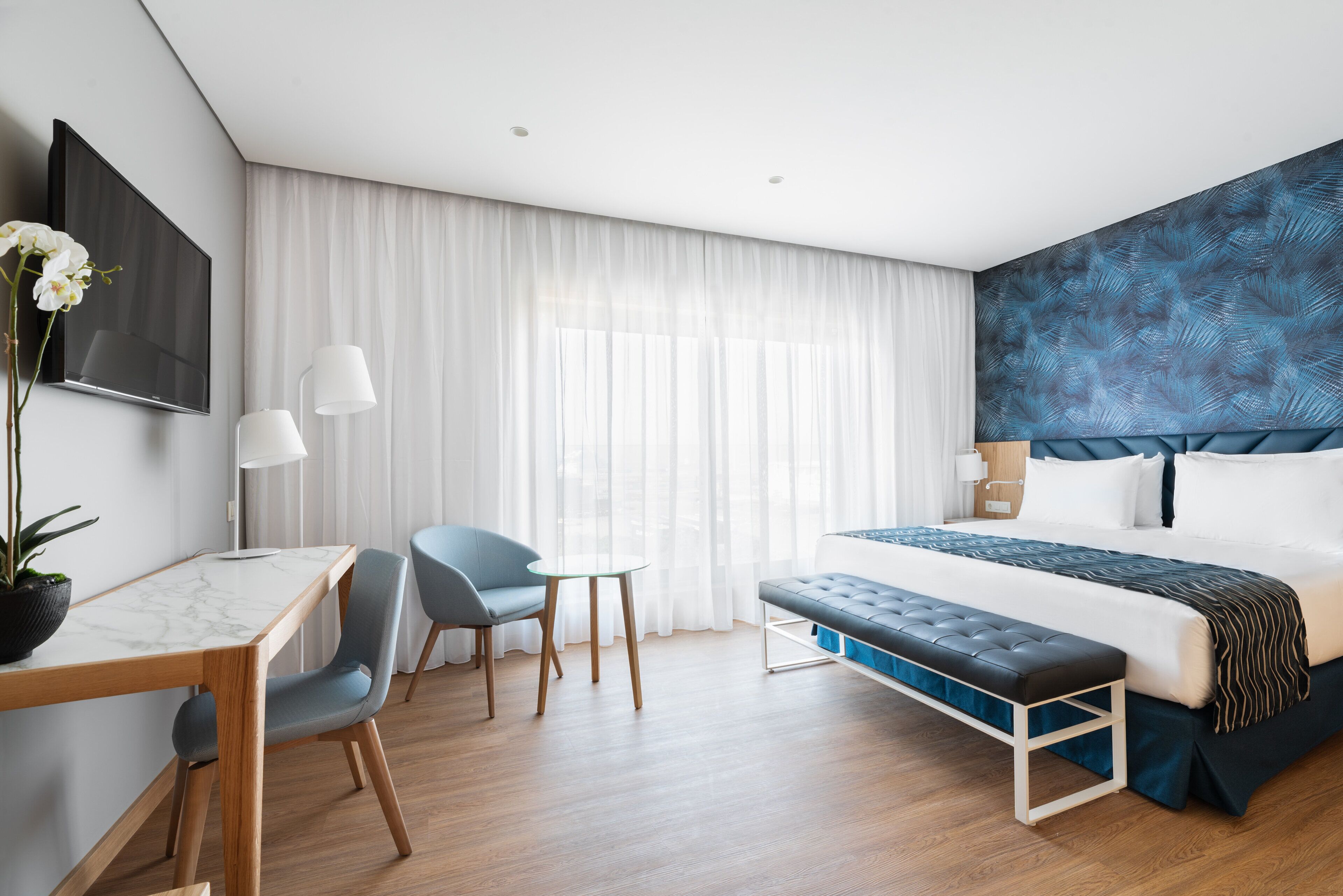 A modern hotel room with a large bed, a desk with a chair, a small table, and a bench.