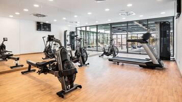 A modern gym with various exercise equipment, large windows, and a wooden floor.