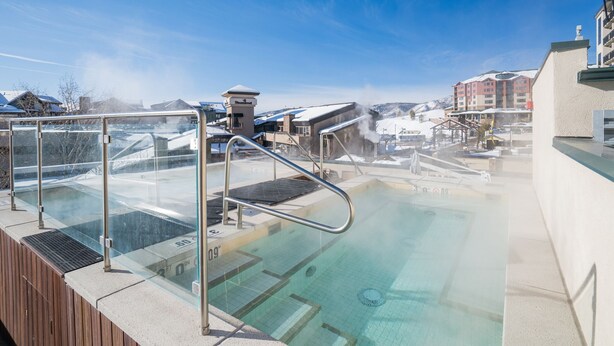 A hot tub area with a glass enclosure, steps leading into the water, and a clear view of the surrounding buildings. - A/C, Pool, Slopeside, Hot Tub, Elevator, Ski Locker, In Unit Washer/Dryer (Steamboat Springs)