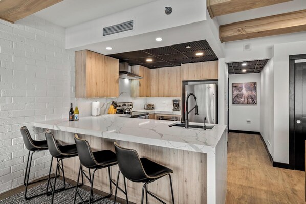 A modern kitchen with a marble island, black bar stools, wooden cabinets, and a stainless steel refrigerator. - Slopeside, Pool, Hot Tub, Elevator, Ski Locker, Washer/Dryer, Ski-In (Steamboat Springs)