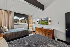 A modern bedroom with two beds, a dresser, a chair, and a large window offering a view of buildings. - Slopeside, Pool, Hot Tub, Elevator, Ski Locker, Washer/Dryer, Ski-In (Steamboat Springs)
