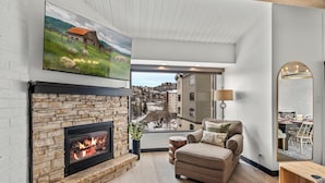 A modern living room with a stone fireplace, a comfortable sofa, and a large window offering a city view. - A/C, Pool, Slopeside, Hot Tub, Elevator, Ski Locker, In Unit Washer/Dryer (Steamboat Springs)