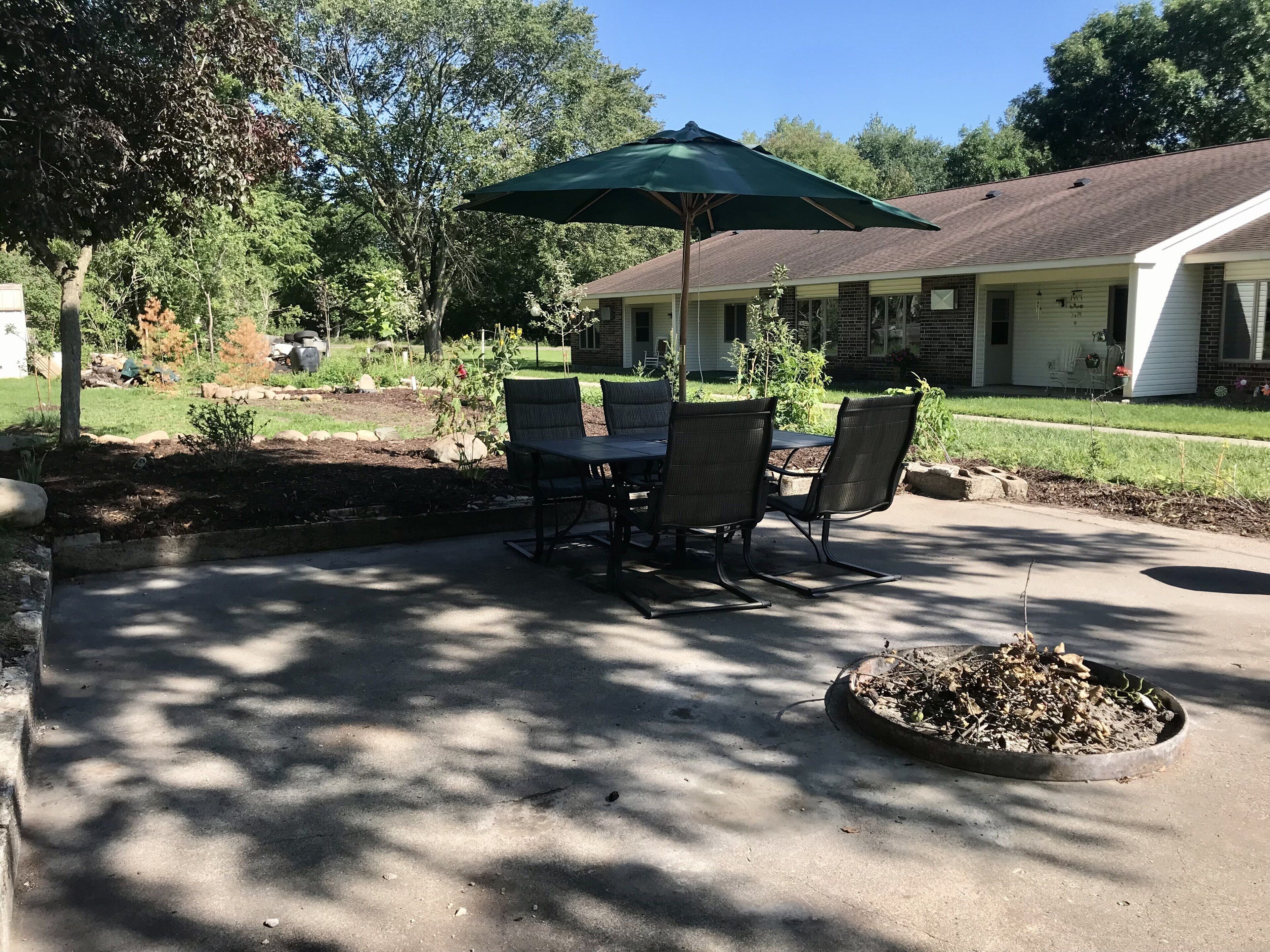 A patio with a table and chairs under a green umbrella, surrounded by a garden and a house in the background.