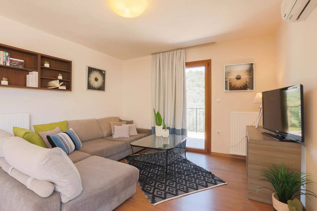 A modern living room with a sofa, coffee table, and TV stand. There are bookshelves and a window with curtains.