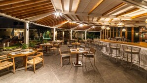 An outdoor dining area with wooden tables and chairs, a bar, and a covered seating area with a view of the garden. - Skiathos Avaton Hotel (Skiathos)