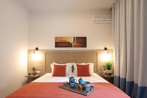 Hotellihuone, jossa on sänky, yöpöydät, seinälle asennettu ilmastointilaite, taulu ja ikkuna, jossa on verhot.