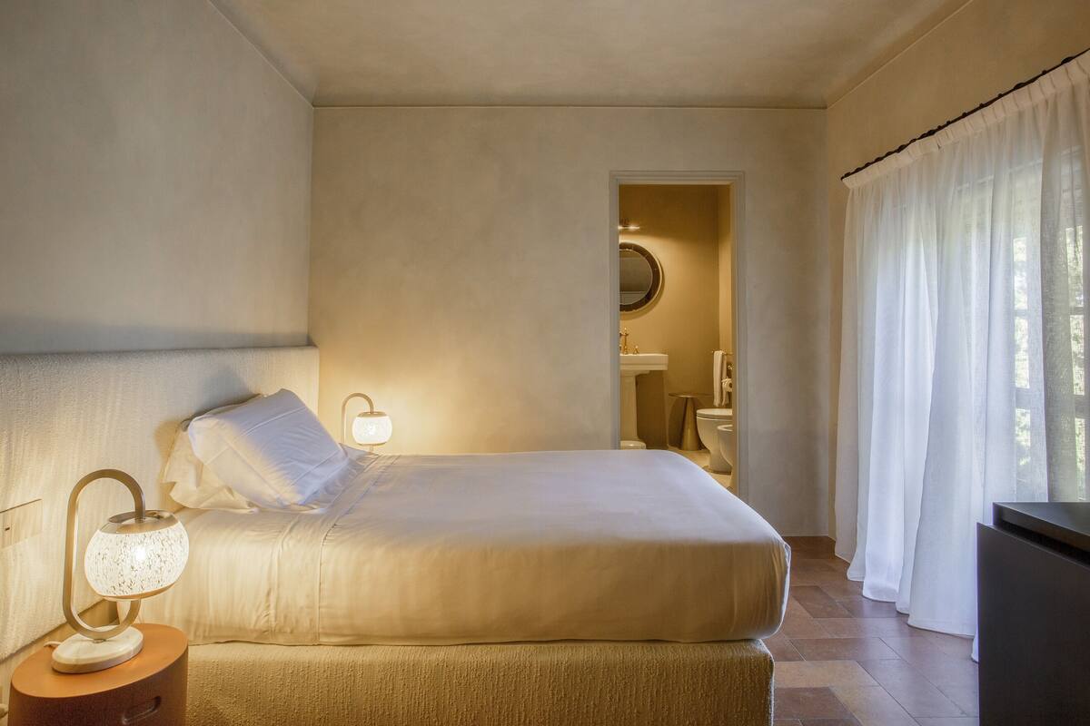 A hotel room with a bed, a bedside lamp, a mirror, a bathroom visible through an open door, and a window with curtains.