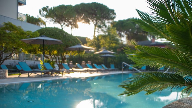 A swimming pool with lounge chairs and umbrellas, surrounded by trees and a building. - Hotel Arizona (Lignano Sabbiadoro)