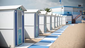 A row of beach huts with a blue and white striped path leading to them, a Swedish flag, and a building with blue and white stripes in the background. - Hotel Arizona (Lignano Sabbiadoro)