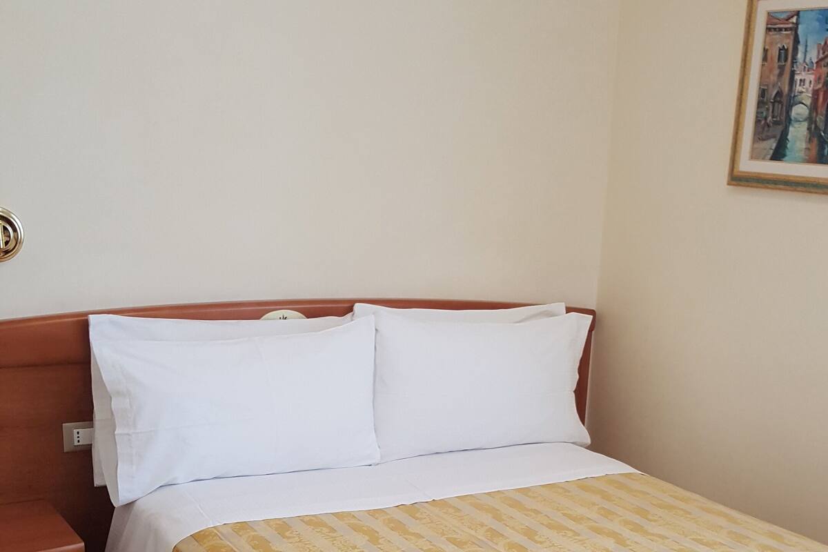 Een hotelkamer met een bed, houten hoofdbord, witte kussens, een gestreept dekbedovertrek, een witte handdoek op het bed, een nachtkastje en een schilderij aan de muur.