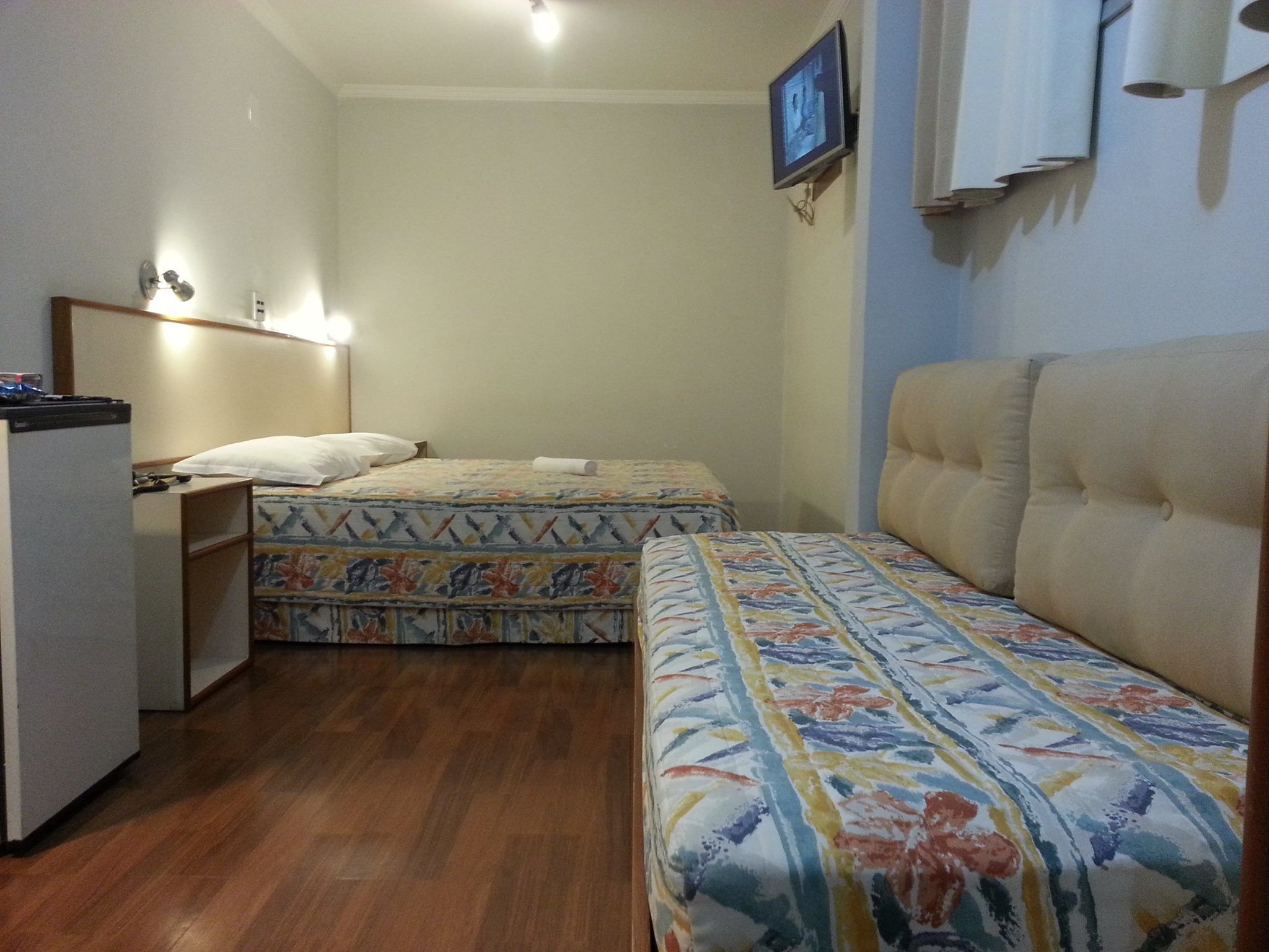 A small, compact room with two beds, a desk, and a television mounted on the wall.