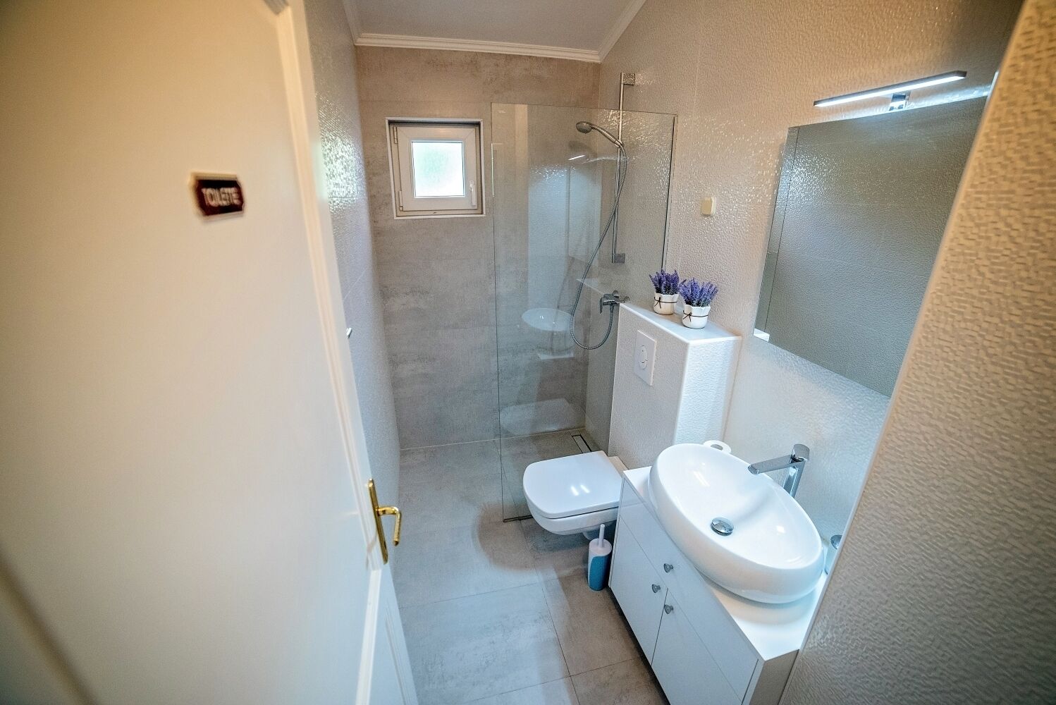 A bathroom with a toilet, sink, and shower. There is a small window above the toilet and a mirror above the sink.
