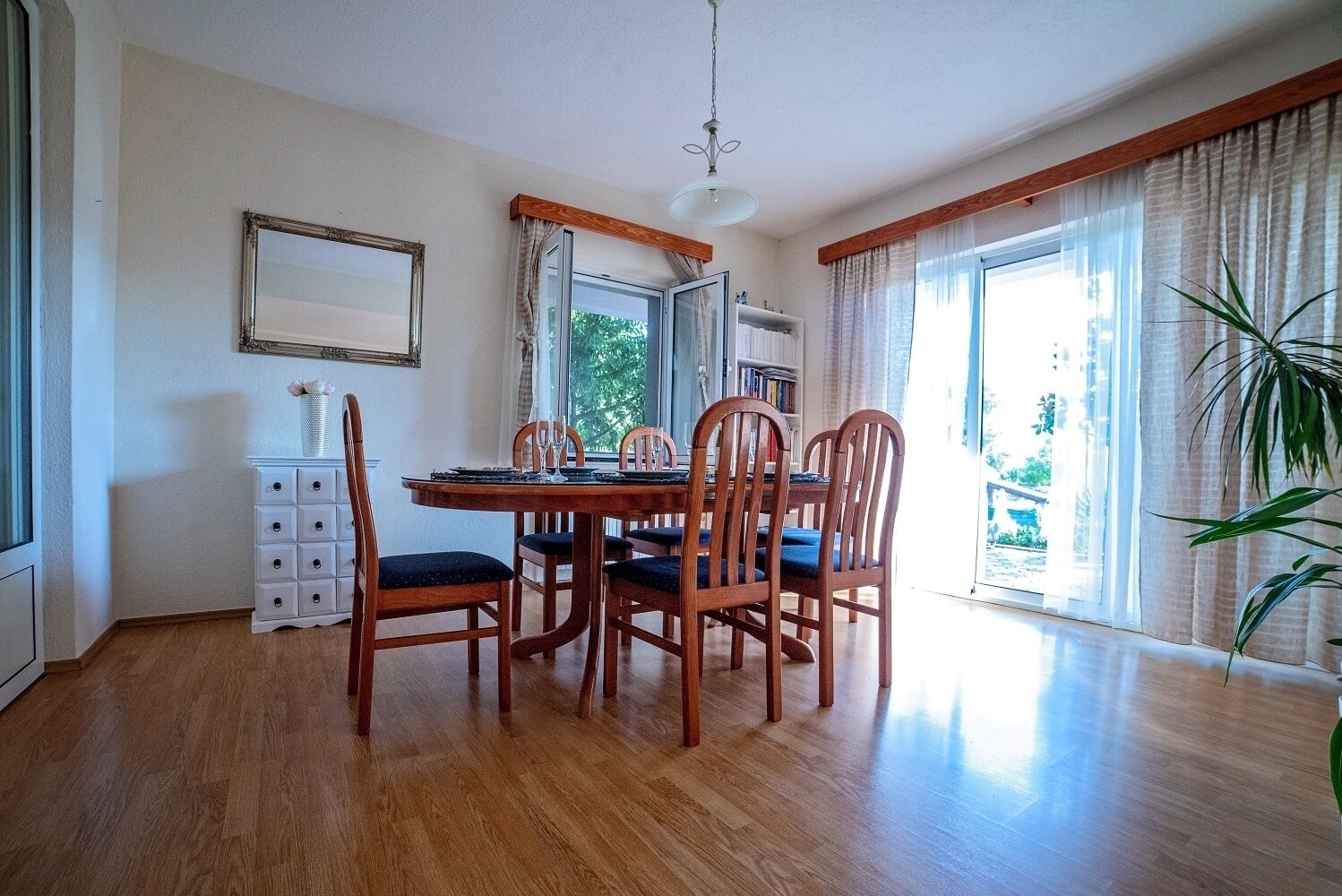 A dining area with a wooden table and chairs, a white cabinet, and a large window with curtains.