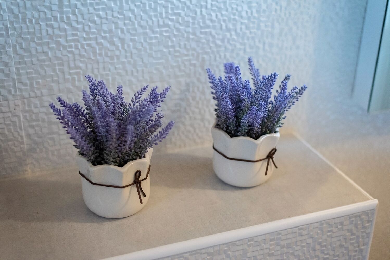 Zwei weiße Keramiktöpfe mit braunen Schleifen, jeweils mit violettem Gr ün bepflanzt, auf einer weißen Oberfläche vor einer strukturierten Wand.