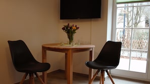 A room with a round wooden table, two black chairs, a vase with flowers, and a TV mounted on the wall. - Ferienwohnung Remise (Lübbenau/Spreewald)