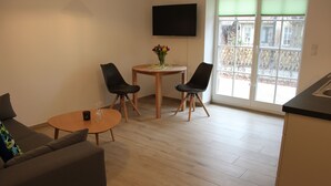 A modern living room with a sofa, a coffee table, two chairs, a small table with a vase of flowers, a flat-screen TV mounted on the wall, and a large window with blinds. - Ferienwohnung Remise (Lübbenau/Spreewald)