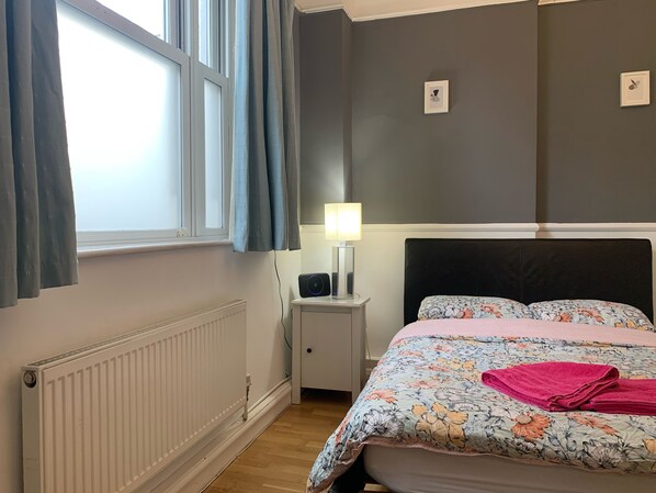 A bedroom with a bed, a bedside table with a lamp, a radiator, and a window with curtains. - Keysan House 1 (London)