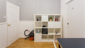 A room with a white door, a white shelving unit, a vacuum cleaner, and a small table. - Keysan House 1 (London)