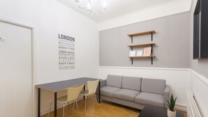 A modern room with a grey sofa, a wooden table, and a desk with two chairs. There is a wall-mounted shelf with framed pictures and a sign that says "LEARN YOUR STREETS". - Keysan House 1 (London)