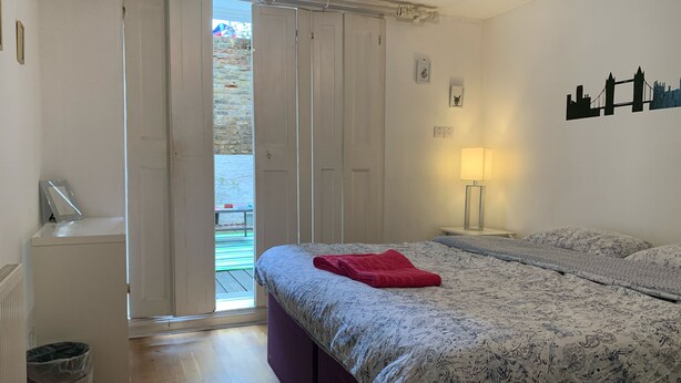 A bedroom with a bed, a nightstand, a lamp, a trash bin, and a sliding door to a balcony. - Keysan House 1 (London)