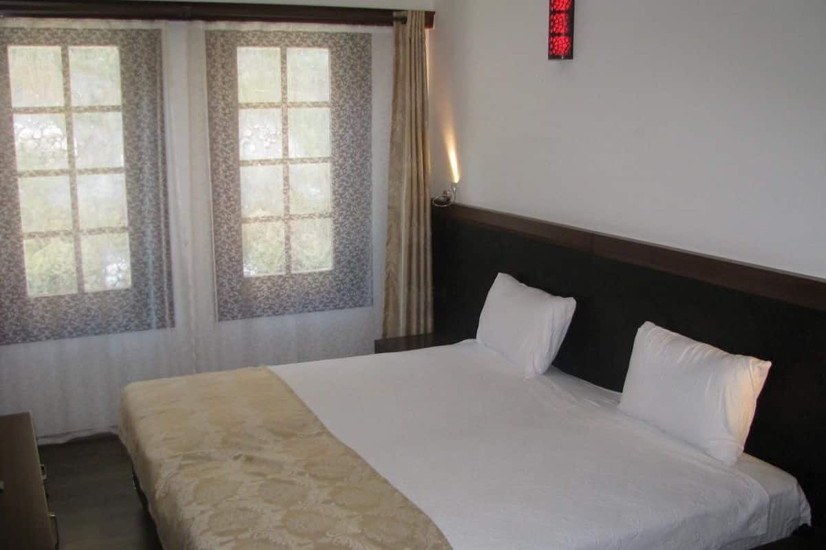 A hotel room with a large bed, two windows with curtains, a bedside table, and a wall-mounted light.