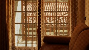 A room with a sofa, a window with lace curtains, and a view of a tiled roof outside.