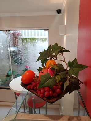 Un comedor con mesa y sillas de vidrio, una cesta de fruta y vistas a un jardín a través de un ventanal.