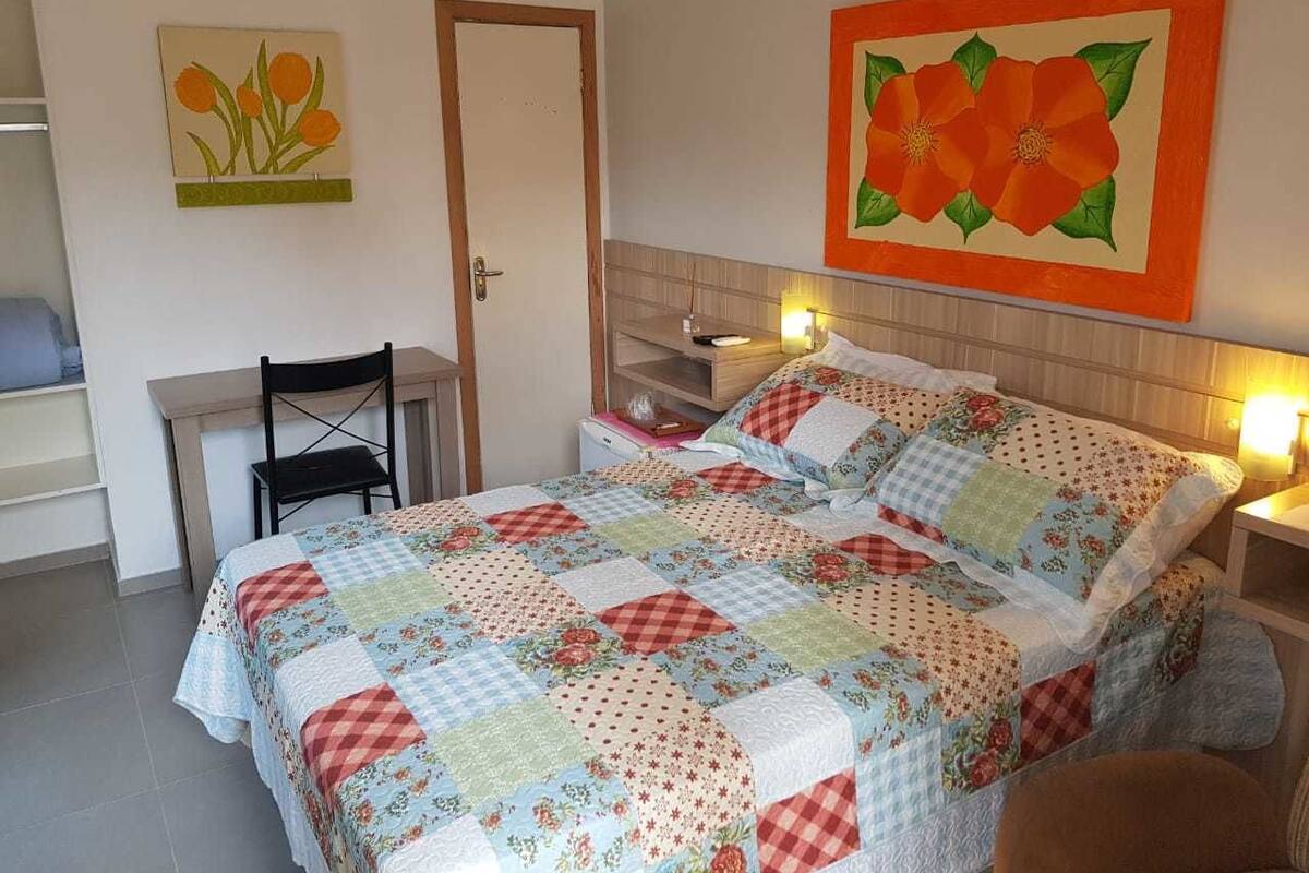 A bedroom with a bed, a desk, and a chair. There are two paintings on the wall.
