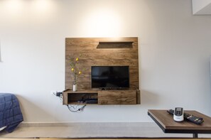 A modern living room with a mounted TV, wooden wall paneling, a small shelf with a vase of flowers, and a coffee table with two remote controls. - Soho Loft Studio con Estilo en Mercedes (Asunción)