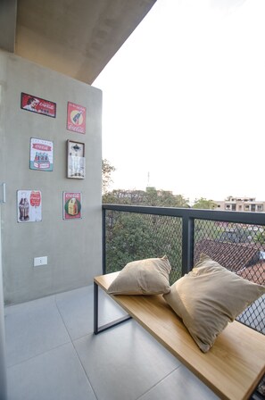 A balcony with a wooden bench and cushions, a concrete wall with vintage Coca-Cola posters, and a view of buildings and trees in the background. - Soho Loft Studio con Estilo en Mercedes (Asunción)