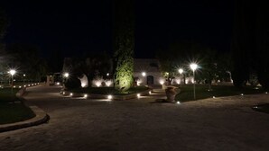 A nighttime view of a courtyard with a building, illuminated by street lamps, surrounded by trees and a central column. - San Marco Antico Relais (Bitonto)