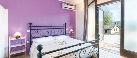 A room with a bed, purple walls, an air conditioning unit, a lamp, and a view through a glass door.