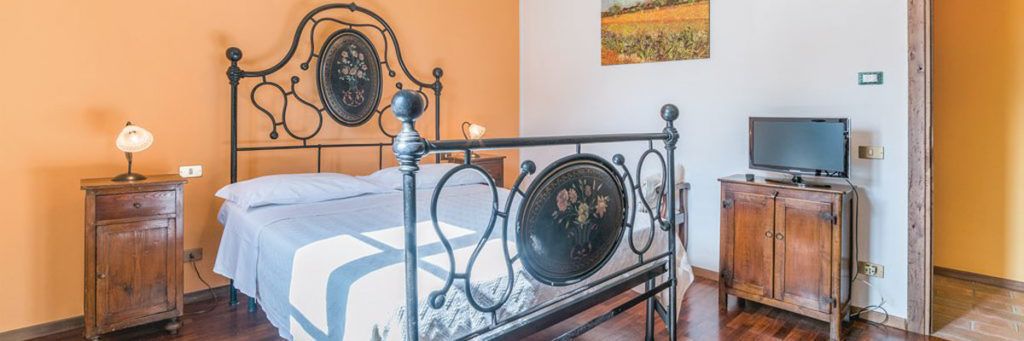 A bedroom with a metal bed frame, a wooden nightstand with a lamp, a television on a wooden cabinet, and a painting on the wall.