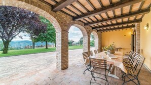 An outdoor patio with a brick floor, wooden tables, and metal chairs. - Casale Villa Chiara (Torgiano)