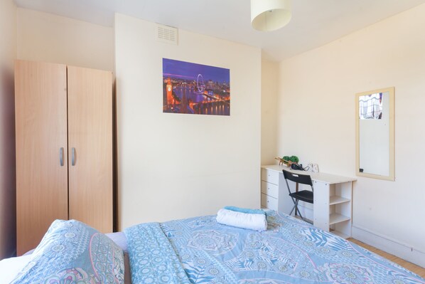 A bedroom with a bed, a desk, a chair, a wardrobe, and a cityscape painting on the wall.