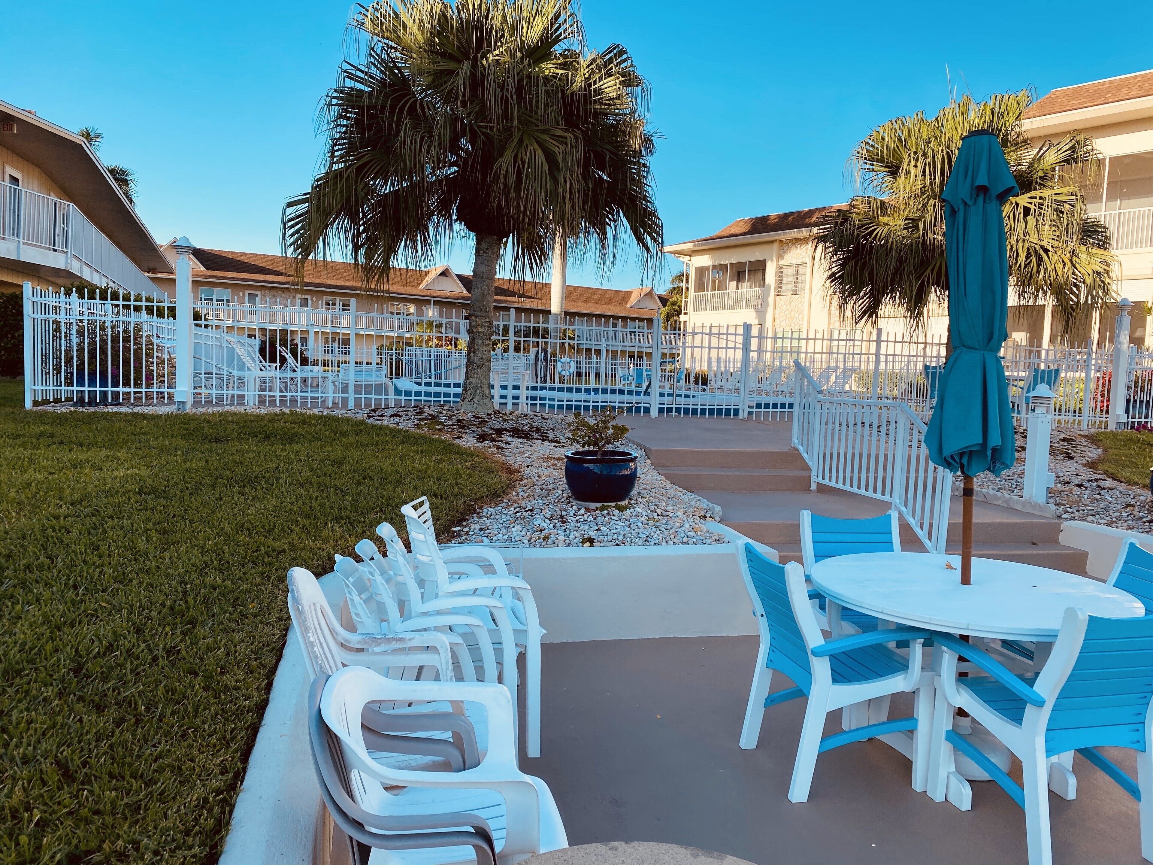 A poolside area with white plastic chairs and a table, surrounded by a white fence, palm trees, and a building with balconies.