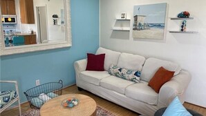 Una sala de estar con sofá, mesa de centro, sillón, espejo y una cocina visible a través de una puerta abierta.