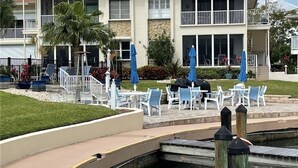 Un edificio de dos pisos con balcón, zona de estar exterior con sillas y sombrillas azules, y una pequeña fuente en la fachada.
