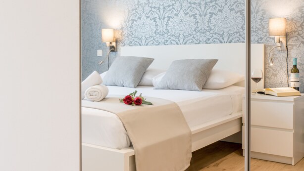 A neatly made bed with white linens and gray pillows, a bedside table with a wine bottle and glass, and a wall with a damask pattern. - Wind Rose Split (Split)