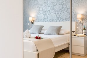 A neatly made bed with white linens and gray pillows, a bedside table with a wine bottle and glass, and a wall with a damask pattern. - Wind Rose Split (Split)