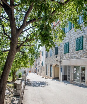 Eine schmale Straße, gesäumt von Steinbauten, einige davon mit grünen Fensterläden, und ein Baum im Vordergrund.