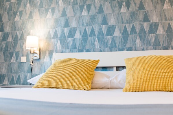 A hotel room with a geometric patterned wall, a white bed with two yellow pillows, and a wall-mounted light fixture. - Wind Rose Split (Split)