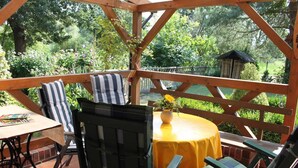 A covered patio with wooden furniture, a table with a yellow tablecloth, and a small garden area with a wooden structure.