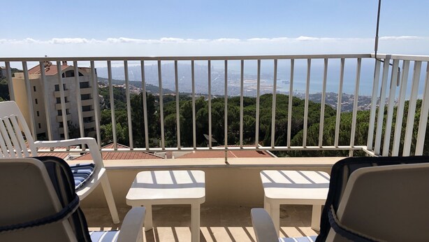 A balcony with white railings, two white chairs with blue cushions, and a view of a cityscape with buildings and greenery. - Splendid Valley & Sea View (Broumana)