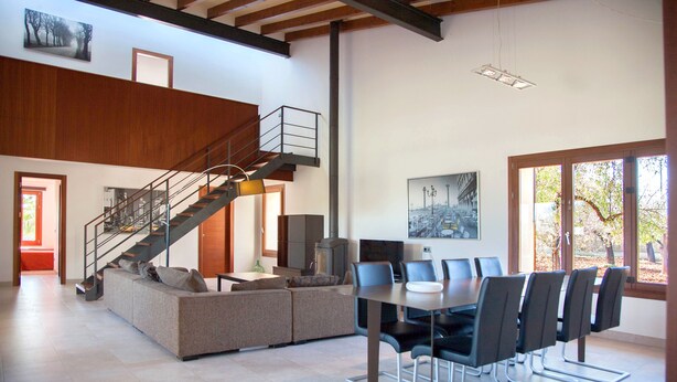 A spacious living area with a large sofa, a dining table with chairs, a staircase, and a large window with a view of trees. - Modern house with large pool  (Búger)