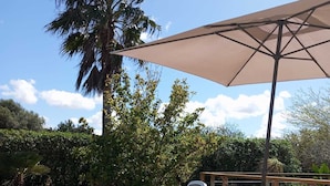 Two white lounge chairs with a beige canopy umbrella, positioned on a tiled patio with a palm tree and other greenery in the background. - Modern house with large pool  (Búger)