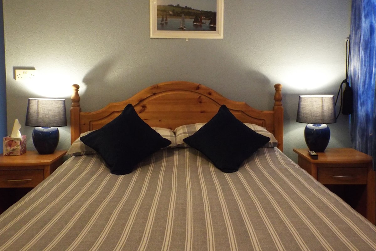 A bed with striped bedding, two black pillows, wooden headboard, two bedside tables with lamps, a framed picture on the wall, and a blue curtain.