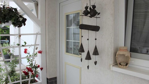 A white door with a window, a hanging wind chime, and a potted plant with red flowers. - Ferienhaus Windspiel (Lübbenau/Spreewald)