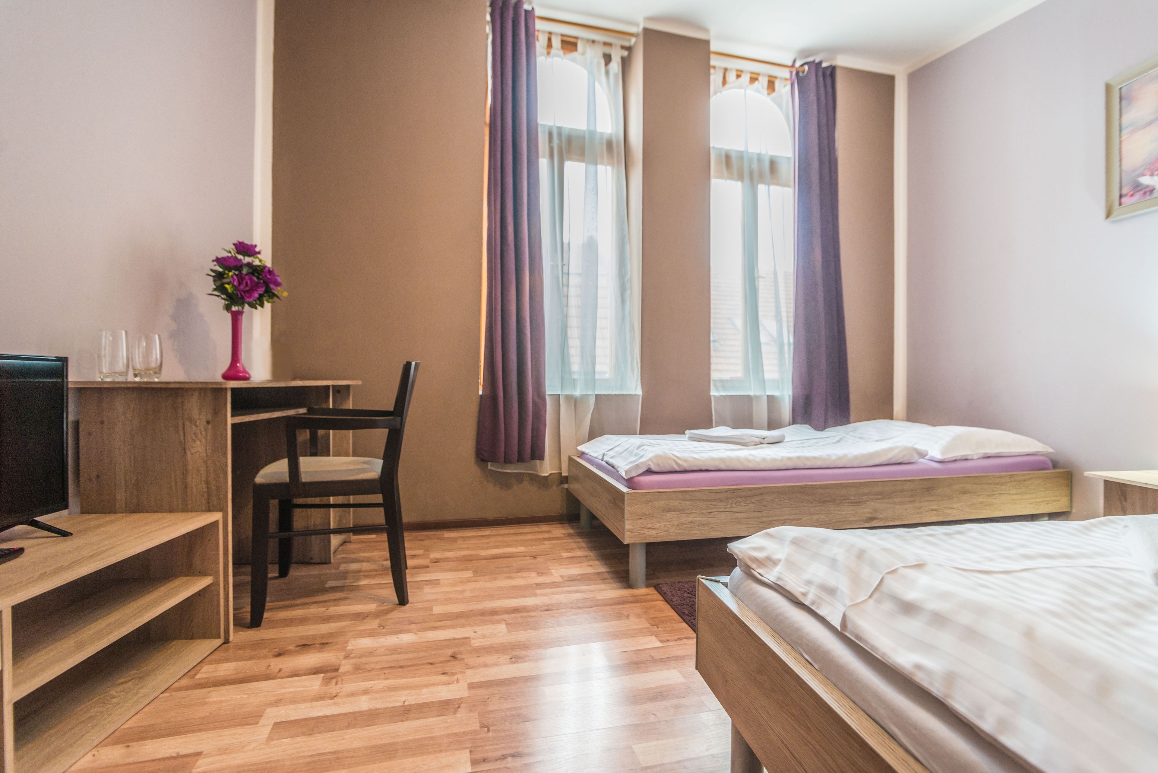 Quarto com cama de madeira, escrivaninha com cadeira, televisão e janela com cortinas.